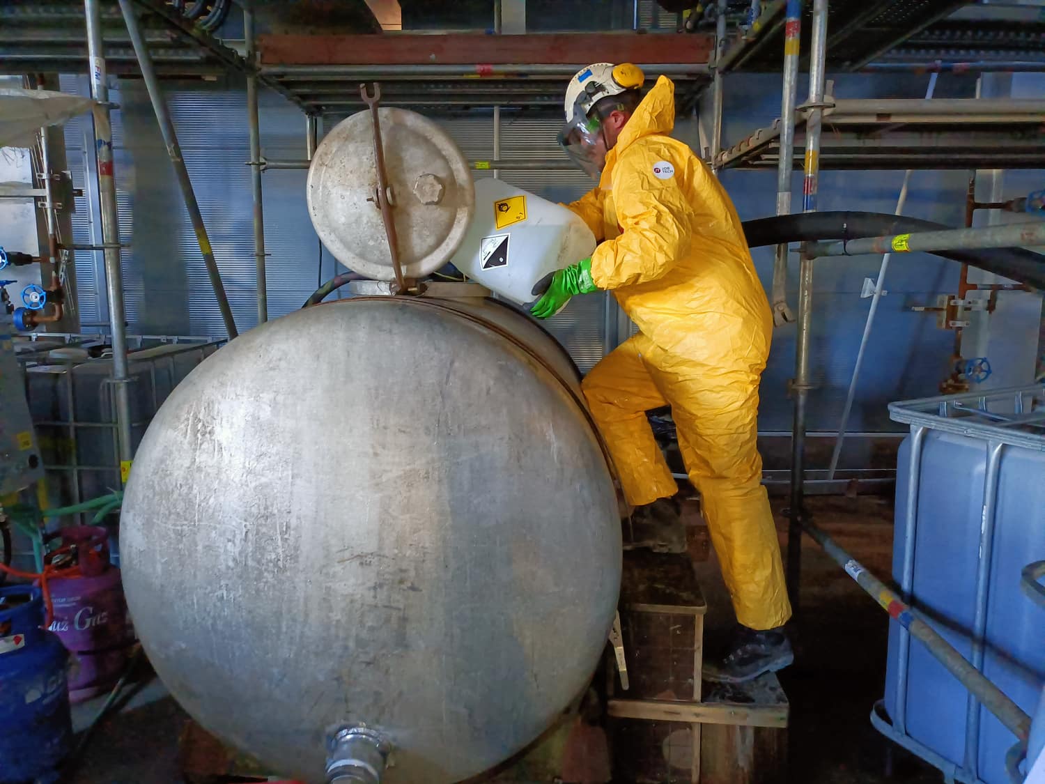 JobTech technician in chemical protective clothing dosing cleaning chemicals into a mixing tank during pipeline chemical cleaning